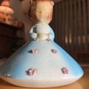 Könnte beinhalten: Eine Keramikfigur mit braunem Haar, die ein hellblaues Kleid mit weißer Basis und kleinen rosa Blumenakzenten trägt. Die Oberfläche der Figur weist durchgehend eine deutlich sichtbare Craquelé-Glasur auf.