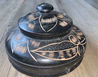 Può includere: Una scatola rotonda di legno nero con coperchio. La scatola e il coperchio sono adornati con disegni floreali e di foglie intagliati. Il coperchio ha un piccolo pomello arrotondato. La scatola è su una superficie di legno.