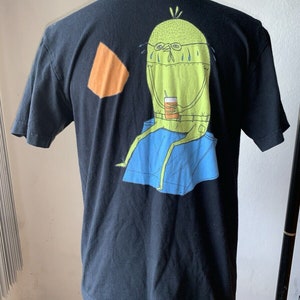 May include: Black t-shirt featuring a cartoon character with glasses, a drink, and blue pants. An orange geometric shape is on the left shoulder area. The graphic is in shades of green, blue, and orange.