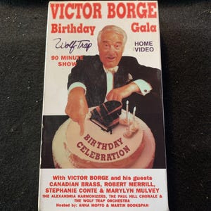 Puede incluir: Una cinta VHS para el Victor Borge Birthday Gala Home Video. La portada presenta a Victor Borge señalando una tarta de cumpleaños con un piano. El texto incluye "Birthday Celebration" y los nombres de los invitados.