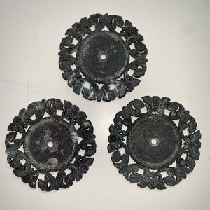 May include: Three antique, dark bronze-colored decorative metal plates. Each plate features a central circular area with a hole, surrounded by a detailed floral and leaf design. The plates have a weathered appearance, suggesting age and use.