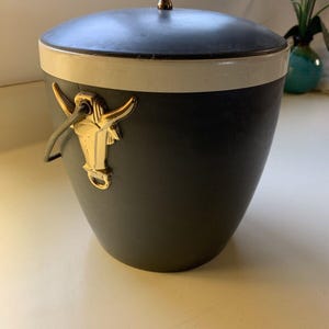 Pode incluir: Um balde de gelo vintage com corpo cinza escuro, faixa branca e tampa preta. O balde apresenta uma alça dourada em forma de cabeça de touro e uma alça dourada na tampa. Uma adição elegante para qualquer bar ou espaço de entretenimento.