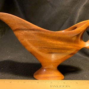 May include: A handcrafted wooden gravy boat with a rich, warm brown hue. The boat features a curved spout, a handle with an open design, and a pedestal base. The wood grain is visible, adding to its natural aesthetic.