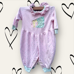 May include: Pink baby sleepsuit with a blue and green embroidered design of a girl holding a gift box. The sleepsuit has a frilled collar and a star pattern.