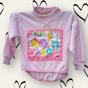 May include: Pink and white striped long-sleeved shirt with a colourful graphic of animals and the word "KAZOO" on the front.