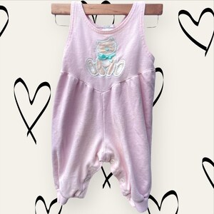 May include: A pink baby onesie with a cute embroidered cat design. The onesie has a sleeveless design and poppers at the bottom.