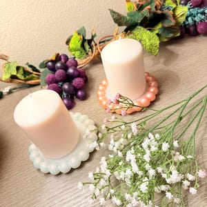 May include: Two white pillar candles on pearl-like candle holders. The candle holders are in different colors, one is pink and the other is light blue. A bouquet of white baby's breath flowers is in the foreground.