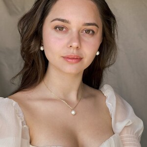 May include: A woman wearing a white, sheer, off-the-shoulder top with pearl earrings and a pearl necklace.