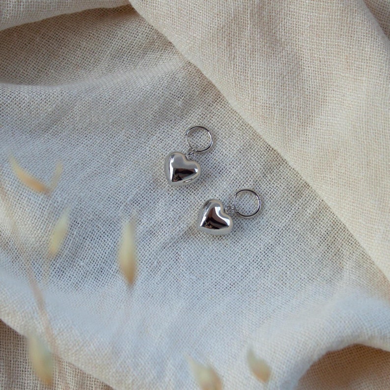 Puede incluir: Dos encantos de plata en forma de coraz&oacute;n con anillos de salto. Los encantos est&aacute;n sobre un fondo de tela beige claro.