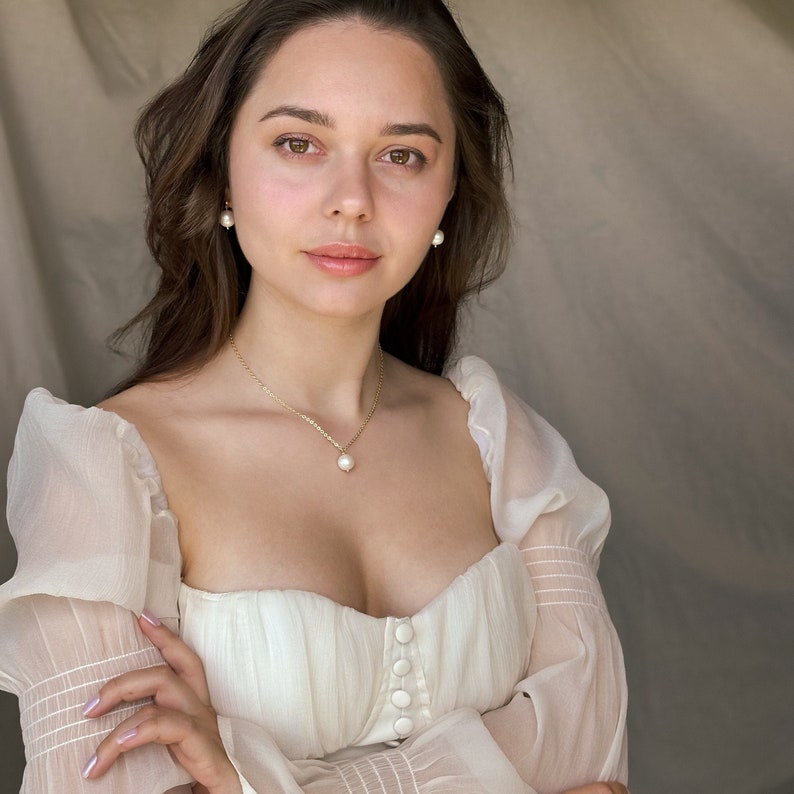 May include: A woman wearing a white, sheer, off-the-shoulder top with pearl earrings and a pearl necklace.