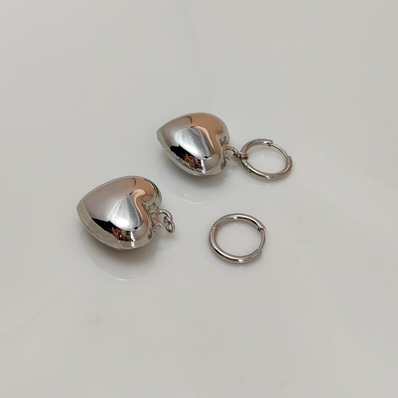 Puede incluir: Un par de pendientes de coraz&oacute;n plateados con cierre de aro. Los pendientes est&aacute;n hechos de metal y tienen un acabado brillante.