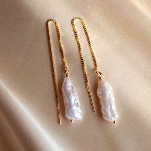 Puede incluir: Un par de pendientes de cadena dorada con una perla barroca blanca colgando de cada cadena.