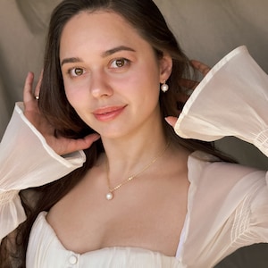May include: A woman wearing a white sheer blouse with a pearl necklace and pearl earrings. The necklace has a single pearl pendant.