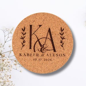 May include: A round cork coaster featuring a dark brown laser-engraved design. The center displays the initials KA with an ampersand, flanked by two leaf sprigs. Below, the text reads KABEER & ALYSON and the date 10.17.2026 in a serif font.