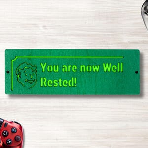 May include: A green wooden sign with the text "You are now Well Rested!" in neon green. The sign features a cartoon character and is flanked by a black and a red game controller, suggesting a gaming theme.
