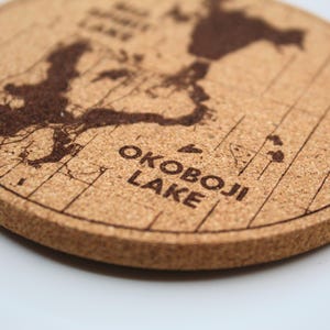 May include: Round cork coaster with a map design. The coaster features a detailed outline of a lake, with the words "OKOBOJI LAKE" printed in a bold font. The coaster is a natural brown color.