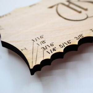 May include: A wooden craft tool with a scalloped edge and black trim. The tool has etched markings for measurements in inches, including 1/16", 1/8", 3/16", 1/4", 5/16", 3/8", and 7/16". The tool also features a decorative design.