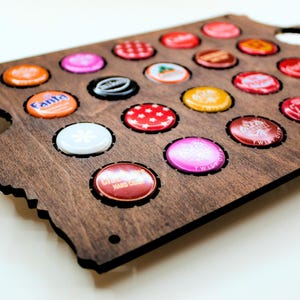 May include: A wooden bottle cap display in the shape of a state, stained dark brown. The display holds various colorful bottle caps. Some caps have text, and others have patterns. The display has a few empty spaces for additional caps.