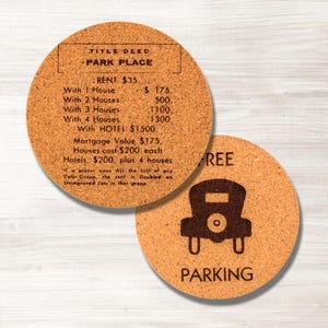 May include: Two round cork coasters. One coaster features text from a board game, including "PARK PLACE" and rent values. The other coaster has a car graphic and the word "FREE PARKING".