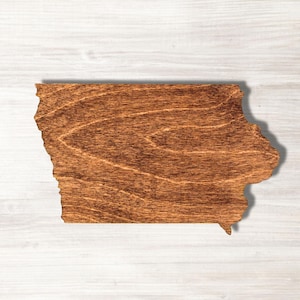 May include: A wooden cutout in the shape of the state of Iowa. The wood has a warm, brown stain, and the grain is visible. The cutout is placed on a white, wood-grain surface.
