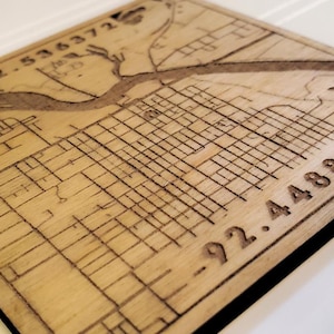 May include: A square wooden map with etched lines depicting a city layout. The map is light brown with dark brown etched details, including streets, buildings, and a river. Coordinates 42.536372 and -92.448826 are visible.