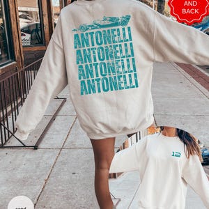 Pu&ograve; includere: Una felpa girocollo color sabbia con una grafica color ottanio sul retro che presenta un'illustrazione di un'auto da corsa sopra il testo ripetuto ANTONELLI. La parte anteriore della felpa mostra il numero 12 in ottanio sul petto.