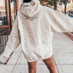May include: A cream-coloured hoodie with a black graphic of the Formula 1 2023 race tracks and flags.