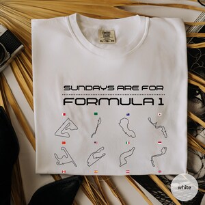 May include: White crew neck sweatshirt with the text "SUNDAYS ARE FOR FORMULA 1" in black. Below the text are black outlines of race tracks with corresponding flags. The sweatshirt is a Comfort Colors brand.