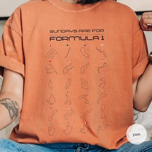 May include: Orange t-shirt with the text "SUNDAYS ARE FOR FORMULA 1" in black. The shirt features black outlines of Formula 1 race tracks with small flags. The t-shirt is a solid orange colour.