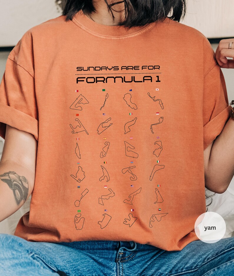 May include: Orange t-shirt with the text "SUNDAYS ARE FOR FORMULA 1" in black. The shirt features black outlines of Formula 1 race tracks with small flags. The t-shirt is a solid orange colour.