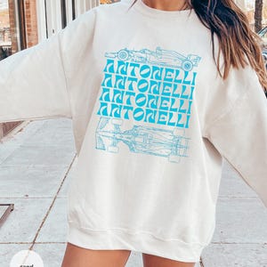 May include: A sand-coloured crewneck sweatshirt featuring a bright blue graphic print. The design includes a top and bottom view of a Formula 1 race car with the name ANTONELLI repeated four times in a stylised, wavy blue font in the centre.
