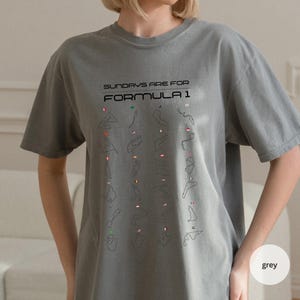 May include: Grey t-shirt featuring the text "SUNDAYS ARE FOR FORMULA 1" and a grid of race track outlines with flags. The shirt is a solid grey colour and has short sleeves.