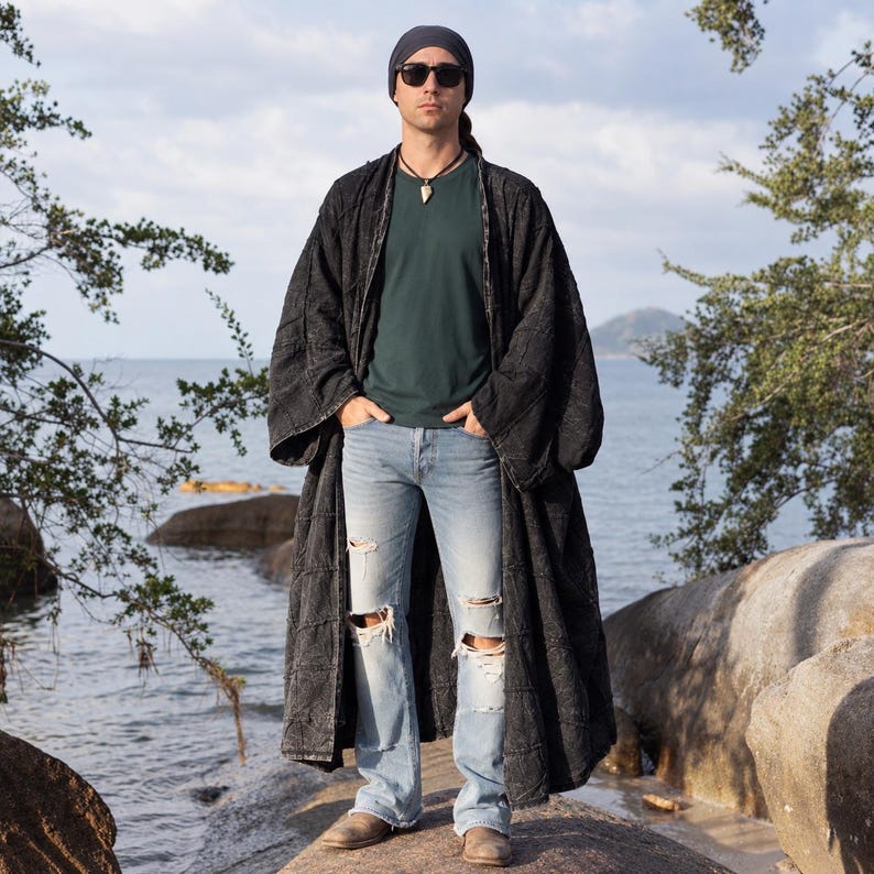 Men’s Akar patchwork kimono duster by MarxSilver shown full length on a coastal rock setting. The model wears the oversized black stonewashed cotton duster open over a deep green shirt, distressed light-wash jeans, boots, sunglasses, and a dark head wrap. The longline silhouette, wide relaxed sleeves, patchwork texture, open front, and dramatic ankle-length proportions are clearly visible against the ocean backdrop.