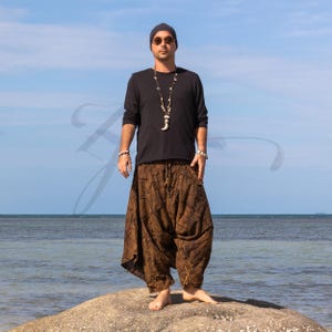 Peut inclure: Une personne porte un haut noir à manches longues et un pantalon bouffant marron sur un rocher près de l'océan. Le pantalon a un motif texturé. La personne porte un bonnet noir, des lunettes de soleil et un collier avec un pendentif.