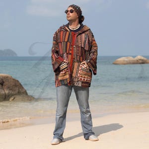 May include: A man wearing a multi-colored patchwork jacket with a hood, blue jeans, and sunglasses. He is standing on a sandy beach.