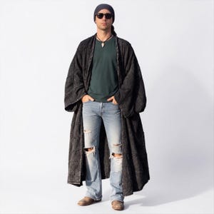 Full-length studio view of the MarxSilver Akar men’s black patchwork kimono duster on a front-facing model. The neutral background clearly shows the oversized proportions, open front, wide relaxed sleeves, ankle-length silhouette, and generous drape. The model wears a dark green shirt, distressed light jeans, boots, sunglasses, and a head wrap, allowing the stonewashed black cotton patchwork texture and handcrafted construction to remain the focus.