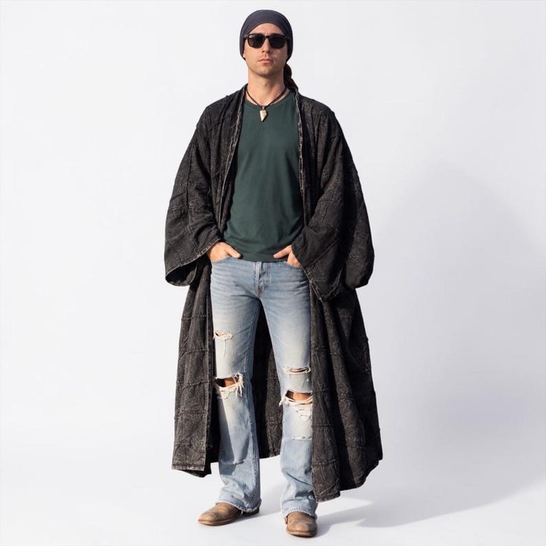 Full-length studio view of the MarxSilver Akar men’s black patchwork kimono duster on a front-facing model. The neutral background clearly shows the oversized proportions, open front, wide relaxed sleeves, ankle-length silhouette, and generous drape. The model wears a dark green shirt, distressed light jeans, boots, sunglasses, and a head wrap, allowing the stonewashed black cotton patchwork texture and handcrafted construction to remain the focus.