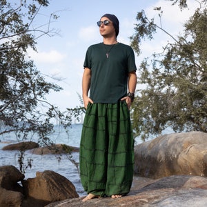 Peut inclure: Un homme portant un t-shirt vert foncé et un pantalon vert foncé à carreaux se tient sur une plage rocheuse. Il porte des lunettes de soleil et un bonnet bleu foncé.