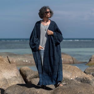 May include: A long, dark blue, open-front robe featuring a textured, grid-like stitched pattern throughout the fabric. The garment has wide, loose sleeves and a straight hemline, worn over a light gray tank top and blue denim jeans.