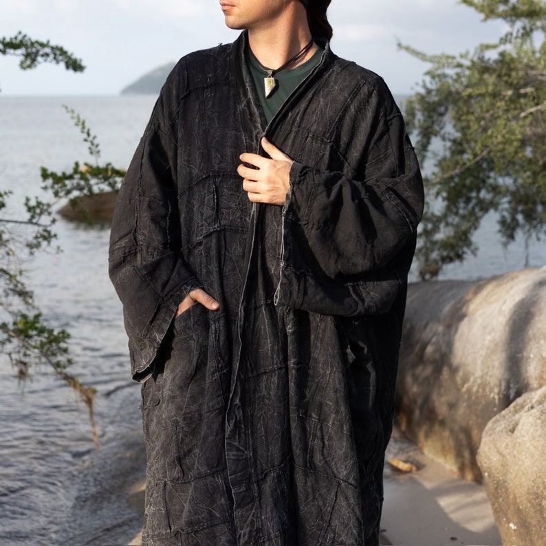 Closer front view of the MarxSilver Akar black patchwork kimono duster, showing the textured stonewashed cotton and handcrafted panel construction. The model holds the open front while one hand rests near the pocket, revealing the oversized sleeve shape and relaxed drape. Subtle tonal stitching, irregular patchwork seams, and the soft worn surface give the long men’s coat a distinctive artisan and modern bohemian character.