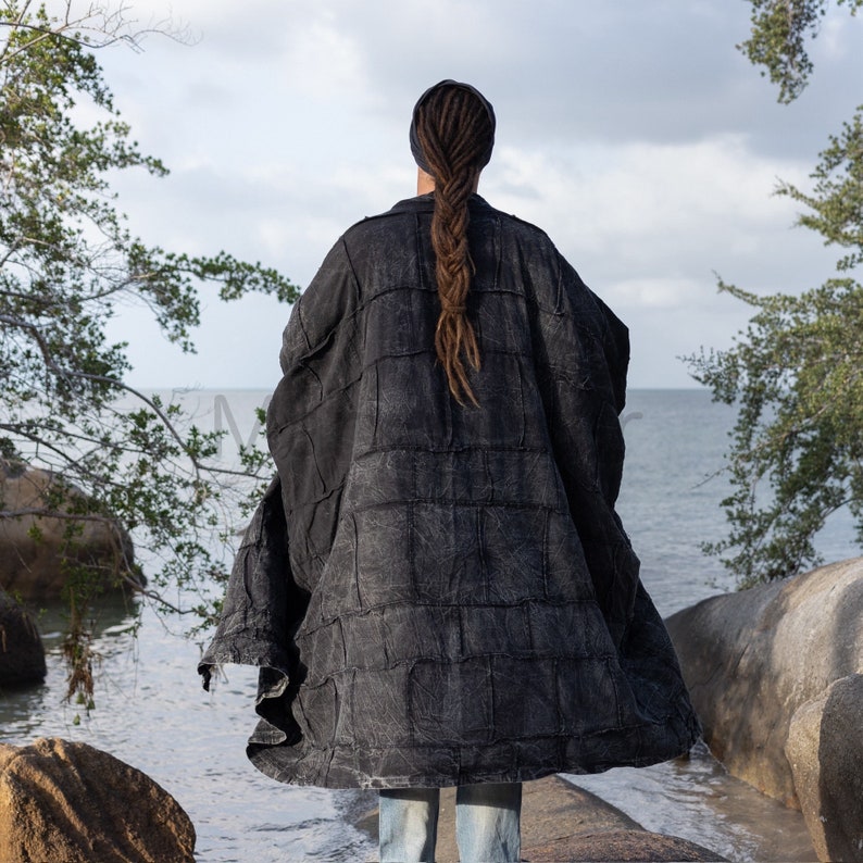 Full back view of the Akar oversized men’s kimono duster in black stonewashed cotton. Large rectangular patchwork panels and exposed artisan-style seams create a textured boro-inspired surface from shoulder to hem. The relaxed coat has wide sleeves, generous proportions, and a long sweeping silhouette. Photographed against rocks and ocean scenery, the image highlights the dramatic scale and handcrafted construction of this MarxSilver design.