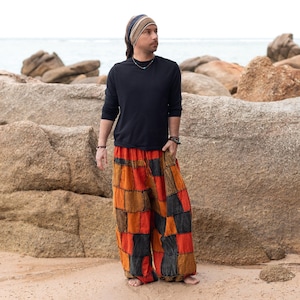 May include: A person wearing a black long-sleeve shirt and patchwork pants in shades of orange, brown, and black. The pants have a wide-leg design. A striped headband and jewelry complete the look. The background features rocks and the ocean.