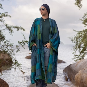 May include: A long, patchwork kimono in teal and green hues. The kimono has a relaxed fit with pockets and is worn over a dark green top and jeans. The person is wearing a dark beanie and sunglasses.