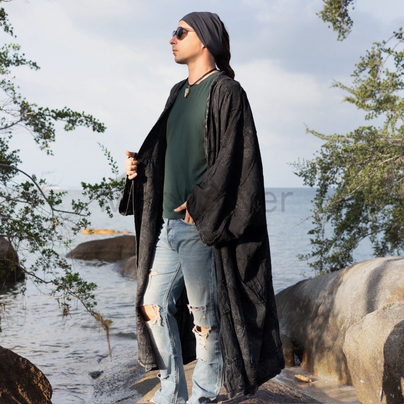 Side-profile view of the Akar men’s black patchwork kimono duster by MarxSilver. The model wears the long 100% cotton coat open over a dark green shirt and distressed jeans on a rocky coastline. Oversized drop shoulders, generous sleeves, relaxed drape, front pocket placement, and the long flowing hem are visible. Stonewashed patchwork panels create subtle texture and an artisan, contemporary bohemian look.