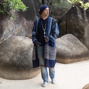 Może przedstawiać: Długa, indygowa kurtka w stylu kimono z wzorzystymi wykończeniami, noszona na ciemnoniebieskiej koszuli i podartych dżinsach. Kurtka ma geometryczny wzór na obszyciu i rękawach. Niebieska czapka i naszyjnik dopełniają stylizację.