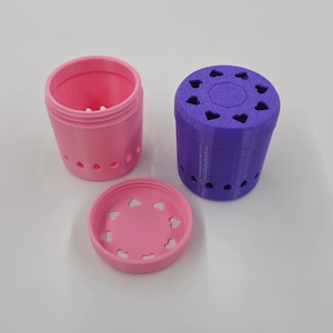 May include: A pink and purple container set with heart-shaped cutouts. The set includes a pink container with a lid and a purple container. The containers are cylindrical and appear to be 3D-printed.