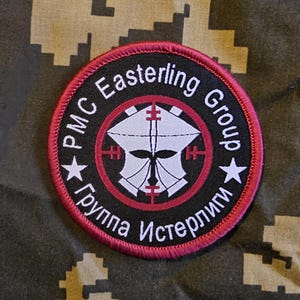May include: A circular patch with a black background and a red border. The patch features the text "PMC Easterling Group" in white, along with a stylized white emblem and two white stars. The text "Группа Истерлинги" is also present.