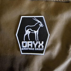 Peut inclure: Écusson brodé noir et blanc avec le mot « ORYX » et le texte « WARFARE GROUP ». L'écusson représente le contour blanc d'un oryx sur fond noir, dans un hexagone bordé de blanc.