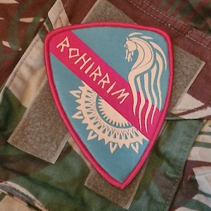 Peut inclure: Un écusson brodé bleu et rose avec un cheval blanc et le mot "ROHIRRIM" en lettres blanches.