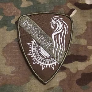 Puede incluir: Un parche bordado en forma de escudo de color marrón oscuro con una franja diagonal verde oliva oscuro. El parche presenta el texto blanco ROHIRRIM, una cabeza de caballo blanca estilizada y un diseño de sol con rayos puntiagudos.
